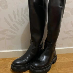 Svarta höga boots med grov sula - Säljer ett par svarta boots med högt skaft och chunky, grovt mönstrad sula. Skorna har rund tå och är tillverkade i blankt skinn som ger en snygg och stilren look. Perfekta för dig som gillar att sticka ut med dina skor och vill ha något som känns både trendigt och robust.