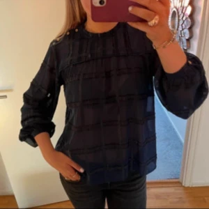 Mörkblå blus med knappar på axeln - Snygg mörkblå blus med subtilt rutigt mönster och dekorativa knappar på ena axeln. Blusen har rund hals och ballongärmar med knappdetalj vid ärmslut. Materialet är lätt och något transparent, perfekt för en cool och trendig look.