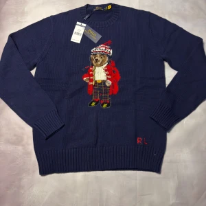 Marinblå stickad tröja Polo Bear - Marinblå stickad tröja från Polo Ralph Lauren med ikoniska Polo Bear broderad framtill. Tröjan har rund hals, ribbade muddar och RL-broderi vid nederkanten. Materialmix av ull, bomull och kashmir för extra mjuk känsla. Perfekt statement-plagg för dig som gillar klassisk stil med twist.