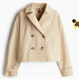 Beige croppad trenchcoat från H&M - Stilren croppad trenchcoat i beige från H&M med dubbelknäppning och klassisk krage. Jackan har långa ärmar med snygga detaljer vid ärmsluten 