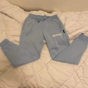 Ljusblå mjukisbyxor från Hoodrich - Säljer ett par ljusblå mjukisbyxor från Hoodrich i storlek M men sitter mer som S. Byxorna har snörning i midjan, ribbade muddar vid bensluten och tryckt logga på ena benet. Mjukt och skönt material, perfekt för chill eller streetwear. Fickor på sidorna.