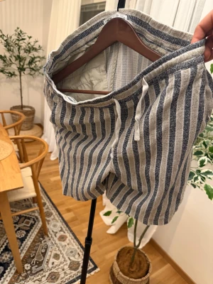 Randiga shorts från Zara - Snygga randiga shorts från Zara i beige och mörkblått. De har en relaxed fit med snörning i midjan och är tillverkade i ett mjukt bomullstyg. Perfekta för varma dagar när du vill ha en chill och stilren look.