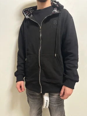 Svart Burberry hoodie med rutig huva - Svart hoodie från Burberry med dragkedja och stor broderad B-logga på bröstet. Insidan av huvan har klassiskt rutigt Burberry-mönster i svart, vitt och rött. Jackan har snörning i huvan och två stora fickor framtill. Perfekt för chill och streetwear.