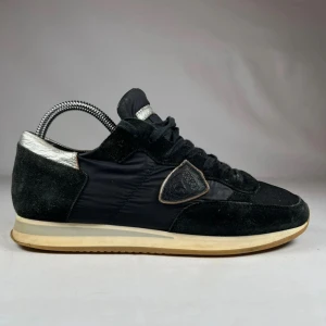 Svarta sneakers från Philippe Model - Snygga svarta sneakers från Philippe Model med detaljer i silver på häl och plös. Ovandel i mocka och textil, med klassisk snörning och rund tå. Beige sula ger en retrovibe. Sköldlogga på sidan för extra look. Pris går att diskutera