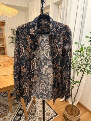 paisleymönstrad skjorta - Snygg skjorta från Morris med paisleymönster och blommiga detaljer i beige och brunt. Skjortan har klassisk krage, knappar framtill och långa ärmar. Perfekt för dig som gillar unika prints och vill sticka ut lite extra.