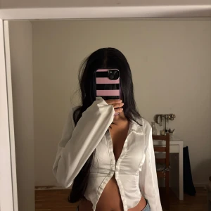 Vit croppad skjorta  - Säljer en snygg vit croppad skjorta från Bershka med lång ärm och snygg krage. Skjortan har knappar framtill och ett smockat parti baktill som ger en cool passform. Materialet är glansigt och känns lyxigt