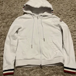 Vit hoodie från Moncler med huva - Snygg vit hoodie från Moncler i bomull med dragkedja framtill och huva med snörning. Tröjan har Moncler-logga på ärmen och ribbade muddar med röd och marinblå rand. Perfekt för dig som gillar stilrena och exklusiva plagg.