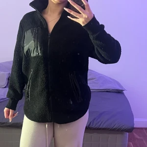 Svart teddyjacka med dragkedja - Mysig svart teddyjacka med hög krage och hel dragkedja framtill. Jackan har två fickor med dragkedja och en cool detalj med en svart syntetficka på bröstet. Perfekt för kalla dagar och chill outfits.