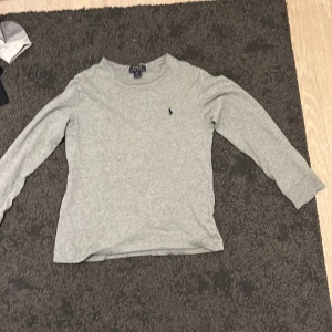 Grå långärmad tröja Polo Ralph Lauren - En stilren grå långärmad tröja från Polo Ralph Lauren i mjuk bomull. Klassisk rund hals och diskret broderad logga på bröstet. Perfekt för dig som gillar enkel och clean stil. Passar dig som vill ha en basic tröja med premiumkänsla.