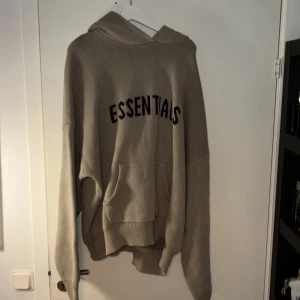 Essentials hoodie stickad XL - Stickad hoodie från Fear of God Essentials i beige med svart logga på ryggen. Oversized fit, huva och känguruficka framtill. Perfekt för dig som gillar streetwear och vill ha en chill vibe. XL men väldigt stor i storleken. 