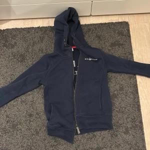 Marinblå hoodie från Sail Racing - Snygg marinblå hoodie från Sail Racing med dragkedja och huva med snörning. Långärmad modell med logga tryckt på bröstet och stort tryck på ryggen. Perfekt för chill dagar och har två fickor framtill. Materialet känns mjukt och är troligen bomull eller polyester.