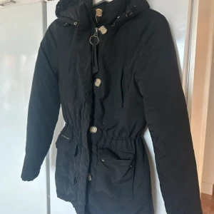 Svart parkajacka från Jeane Blush - Svart parkajacka från Jeane Blush med huva, stora fickor och knappar framtill. Jackan har dragsko i midjan för snygg passform och är långärmad. Perfekt för kyliga dagar och har en stilren look med diskreta detaljer.