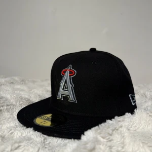 LA Angels svart keps New Era 59FIFTY - Svart LA Angels 59FIFTY keps från New Era med broderad logga i silver och rött framtill, samt jubileumsmärke på sidan. MLB-logga baktill. Platt skärm och klassisk passform. Perfekt för dig som gillar baseball och streetwear.
