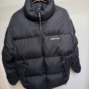 Svart pufferjacka Axel Arigato - Svart pufferjacka från Axel Arigato med hög krage och tydlig logga 'ARIGATO ©2014' på bröstet. Jackan har dragkedja framtill, två sidofickor med dragkedja och är tillverkad i återvunnen polyester. Perfekt för kalla dagar och har en clean, modern look.