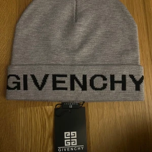  Givenchy mössa - Snygg grå mössa från Givenchy med svart logotyp broderad framtill. Klassisk uppvikt modell i mjukt stickat material som ger en clean och exklusiv look. Perfekt accessoar för att lyfta din outfit med en lyxig touch.