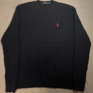 Mörkblå stickad tröja Polo Ralph Lauren - Kabelstickad mörkblå tröja från Polo Ralph Lauren med klassisk rund hals och röd broderad logga på bröstet. Tröjan har ribbade muddar och är långärmad, perfekt för dig som gillar stilren och tidlös design.