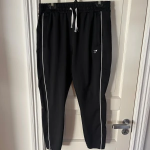 Svarta mjukisbyxor från Gymshark - Svarta mjukisbyxor från Gymshark med vita sidoränder och dragsko i midjan. Byxorna har smal passform, elastisk midja och två fickor framtill. Perfekta för chill eller träning, med diskret Gymshark-logga på låret.