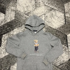 Grå hoodie Polo Bear Ralph Lauren M - Grå hoodie från Polo Ralph Lauren med ikoniskt Polo Bear-tryck på bröstet. Tröjan har huva, ribbade muddar och är tillverkad i mjuk bomullsmix. Perfekt för dig som gillar streetwear och klassisk stil med en twist.