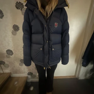 Blå Fjällräven Expedition dunjacka - Säljer en blå Fjällräven Expedition Series dunjacka med huva och gula snören. Jackan har två stora fickor med knappar, dragkedja och tryckknappar framtill samt Fjällräven-logga på bröstet. 