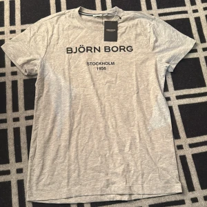 Grå Björn Borg t-shirt med logga - Snygg grå t-shirt från Björn Borg med svart logotyp och texten 'Stockholm 1956' på bröstet. Klassisk rund halsringning och korta ärmar. Tillverkad i mjuk bomull för en skön känsla. Perfekt för en clean och sportig look.