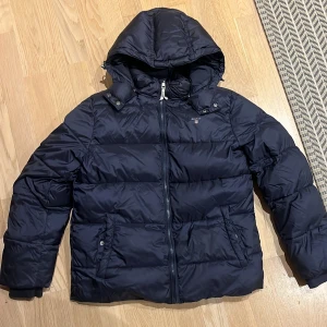 Mörkblå pufferjacka från GANT - Snygg mörkblå pufferjacka från GANT med huva och dragkedja framtill. Jackan har två fickor med knappstängning och ett broderat GANT-logo på bröstet. Insidan har en cool röd detalj och tryckt text. Perfekt för kalla dagar. En lappad defekt men inget som förstör helhetsbilden av jackan eller syns särskilt mycket.