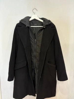 Svart kappa med huva från KappAhl - Svart kappa från KappAhl med snygg huva och dragkedja framtill. Jackan har långa ärmar, två fickor och en stilren look. Yttermaterialet är ullblandning och fodret är i syntet. Perfekt för dig som vill ha en klassisk och varm jacka med modern touch.