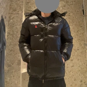 Svart glossy pufferjacka från Polo Ralph Lauren - Svart pufferjacka från Polo Ralph Lauren med röd logga på bröstet. Jackan har huva, dragkedja framtill och två sidofickor med dragkedja. Den är quiltad och har en glansig finish som ger en cool look. Perfekt för kalla dagar.