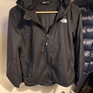 Svart vindjacka från The North Face - Snygg svart vindjacka från The North Face med huva och vita logotyper på bröstet, ryggen och ena ärmen. Jackan har dragkedja framtill och två sidofickor med dragkedja. Perfekt för blåsiga dagar och har en clean, sportig look. 