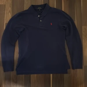 Polo Ralph Lauren pikétröja - Snygg marinblå långärmad pikétröja från Polo Ralph Lauren med klassisk krage och två knappar. Tröjan har det ikoniska röda broderade Polo-logot på bröstet och är gjord i mjuk bomull. Perfekt för dig som gillar stilrena och tidlösa plagg⭐️💫. Hör av dig vid frågor och funderingar🙌
