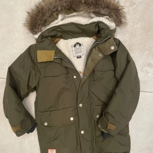 Olivgrön parkajacka från Didriksons - Parkajacka från Didriksons i olivgrönt med fluffig fuskpäls på huvan och mjukt vitt teddyfoder. Perfekt för kalla dagar.