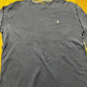 Blå långärmad tröja Polo Ralph Lauren - Snygg blå långärmad tröja från Polo Ralph Lauren i storlek XL. Tröjan är tillverkad i 100% bomull med ett diskret broderat logomärke på bröstet. Den har en klassisk rund hals och en lätt ribbad struktur som ger en cool och avslappnad vibe.