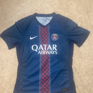 PSG Barcola #29 matchtröja Nike - Säljer en officiell Paris Saint-Germain fotbollströja med Barcola och nummer 29 på ryggen. Tröjan är marinblå med röd och vit randig detalj på framsidan, PSG-logga och Nike Swoosh. Materialet är lätt och andas, perfekt för träning eller match.