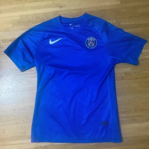 Blå PSG fotbollströja från Nike - Säljer en blå Paris Saint-Germain fotbollströja från Nike med klubbens emblem på bröstet och vit Nike-logga. Tröjan är kortärmad och har en sportig passform i lätt, ventilerande material som passar perfekt för träning eller match.