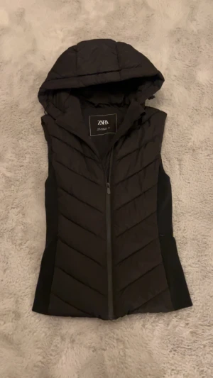 Svart dunväst med huva från Zara - Svart dunväst från Zara med snygg huva och dragkedja framtill. Västen har ett quiltat mönster och är perfekt för lager-på-lager. Stilren design som funkar till många olika outfits. Passformen är normal och materialet är mjukt och lätt. Andvänd 4-5 gånger, köptes för 500kr