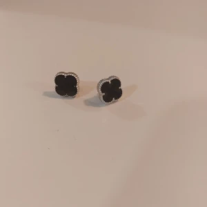 Svarta fyrklöver örhängen i silver - Snygga örhängen med fyrklöverformad design i svart och silver. Kanterna har en detaljerad, pärlad look som ger extra stilpoäng. Perfekt för dig som gillar klassiska smycken med en twist.