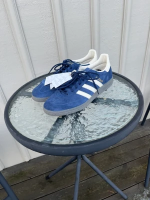 Adidas Spezial blå sneakers mocka - Helt nya, endast provade. Adidas godkännde ej retur. Nypris 1300Säljer ett par Adidas Gazelle sneakers i blå mocka med vita detaljer och klassiska tre ränder på sidan. Skorna har snörning, grå platt sula och vit insida med Adidas-logga. Stilren och retro design som passar perfekt till streetwear. Som även har ett par extra vita snören. 