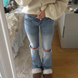 Ljusblå bootcut jeans med slitna knän - Snygga ljusblå jeans med bootcut passform och låg midja. Jeansen har slitna hål vid knäna och coola fickor med lock och knappdetaljer bak. Perfekt för dig som gillar en avslappnad och trendig stil. Lite slitningar längst ner på benet.