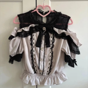 Romantisk blus med spets från Liz Lisa - Supersöt blus från Liz Lisa i ljus rosa med svarta spetsdetaljer och volanger. Blusen har puffärmar, rosett med pärlor och knappar framtill, samt en öppen rygg med knäppning. Perfekt för jirai kei occ andra j-fashion stilar