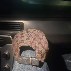 Gucci keps med GG-mönster och läderrem - Snygg beige keps från Gucci med klassiskt GG-monogram i mörkbrunt över hela kepsen. Justerbar rem i svart läder baktill och ikonisk röd-blå rand på sidan. Tillverkad i bomull och polyester med insida i mörkblått tyg. Perfekt för dig som vill ha en lyxig streetstil.