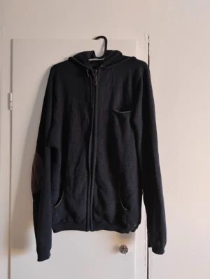 Svart hoodie från Jack & Jones Storlek L - Svart hoodie från Jack & Jones Premium med dragkedja framtill, huva och fickor. Tröjan har förstärkta armbågslappar i mörkare tyg och en liten bröstficka. Perfekt för dig som gillar en enkel och stilren look.