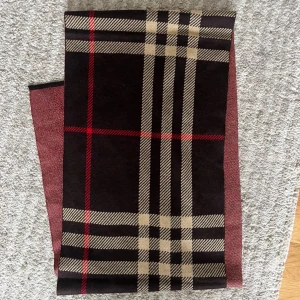 Rutig halsduk i mörka toner - Snygg halsduk med klassiskt rutigt mönster i svart, beige och rött. Perfekt att slänga över axlarna för en cool och tidlös look. Materialet känns mjukt och värmande, och färgkombon gör den enkel att matcha med jackor och tröjor.