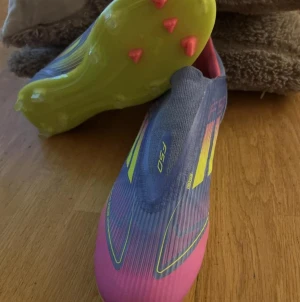 Adidas F50 fotbollsskor coral victory - Säljer ett par Adidas F50 fotbollsskor med en snygg mix av lila, rosa och neon-gul. Skorna har en strömlinjeformad design, platt sula med dobbar och är tillverkade i syntetmaterial för lätthet och snabbhet på planen. Perfekt för dig som vill sticka ut på matchen!