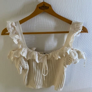 Krämvit croppad topp med volang från Zara - Superfin croppad topp från Zara i krämvit färg. Stickad med ribbad struktur och breda axelband dekorerade med romantiska volanger och spetsdetaljer. Toppen har snörning framtill och är ärmlös, perfekt för att skapa en söt och trendig look.