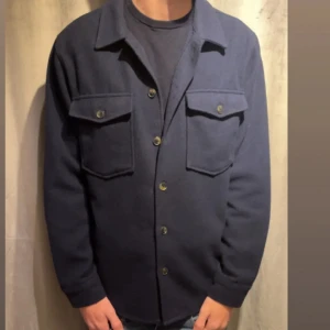 Mörkblå overshirt med fickor - Snygg mörkblå overshirt med klassisk krage och stora bröstfickor med knappar. Skjortan har långa ärmar och stängs med knappar framtill. Perfekt att bära över en t-shirt för en chill och stilren look.