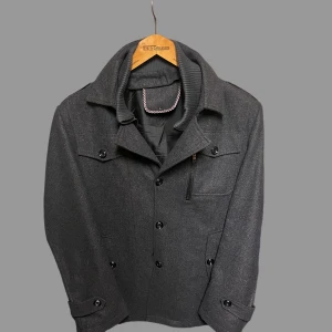 Mörkgrå rock med snygga detaljer - Stilren mörkgrå rock i ullmix med klassisk krage och knappar framtill. Jackan har en bröstficka med dragkedja, två sidofickor med lock och dekorativa knappar vid ärmslut. Insidan har rutigt kantband som ger en cool touch. Perfekt för dig som gillar clean och tidlös stil.