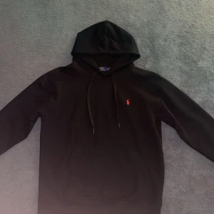 Svart hoodie från Polo Ralph Lauren - Svart hoodie från Polo Ralph Lauren med klassisk röd logobrodyr på bröstet. Tröjan har huva med snörning och en stor magficka framtill. Mjuk bomullskvalitet och långärmad modell, perfekt för chill dagar.
