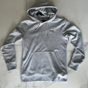 Grå hoodie från Björn Borg - Snygg grå hoodie från Björn Borg med svart logga på bröstet. Tröjan har huva med snörning, stor magficka och ribbade muddar vid ärmslut och nederkant. Perfekt för chill eller träning, enkel och stilren design.