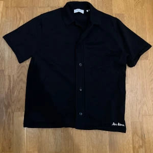  Les Deux  - Svart kortärmad skjorta från Les Deux med broderad logga i vitt vid nederkanten. Skjortan har knappar framtill och ett färgglatt blommigt tryck på ryggen med inslag av blått, rött, gult och grönt. Materialet är mjukt och känns som bomull, perfekt för dig som gillar stilrena detaljer.