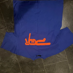 Blå t-shirt med orange tryck PEGADOR - Säljer en blå t-shirt från PGDR med ett stort orange grafiskt tryck på framsidan. T-shirten har rund hals och korta ärmar, och är gjord i mjuk bomull. Passformen är normal och trycket ger en cool streetkänsla.