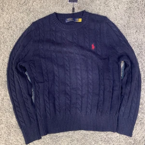 Ralph Lauren cableknit - Kabelstickad marinblå tröja från Polo Ralph Lauren med klassisk röd broderad logga på bröstet. Rund hals, ribbade muddar och långärmad modell. Perfekt för dig som gillar stilrena och tidlösa plagg med premiumkänsla. Den är i storlek S och aldrig använd den sticks inte. 